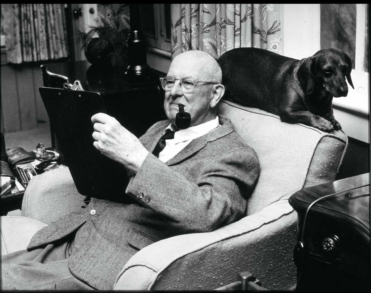 PG Wodehouse sits reading a book and smoking a pipe with a dog perched behind his chair, 1965