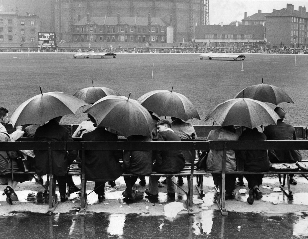 A group of spectators sit underneath umbrellas in the rain, waiting for a match to resume at the Oval, 1946