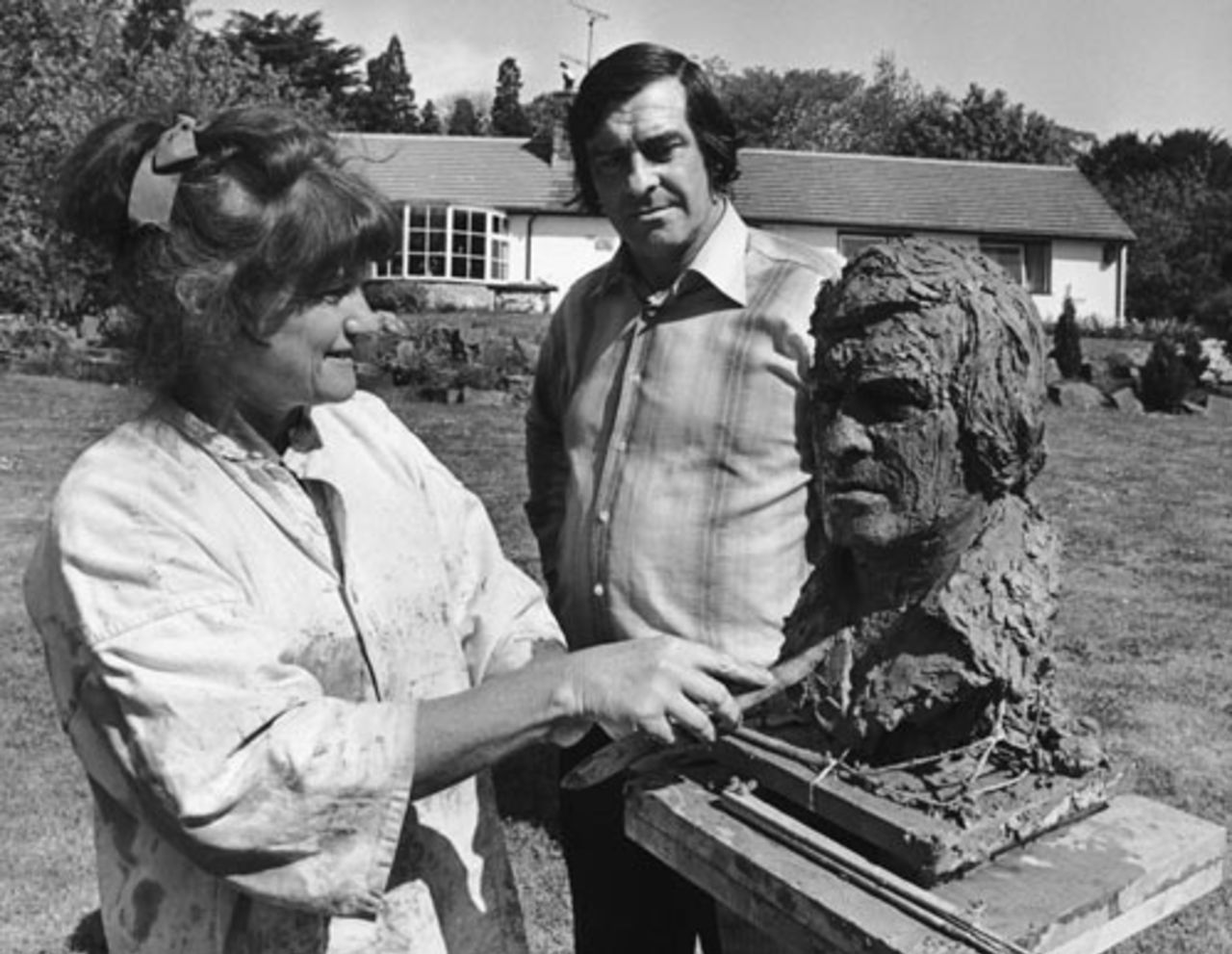 Fred Trueman looks on as Betty Miller works to complete a bust of him in the garden of his Gargrave home, June 10, 1977