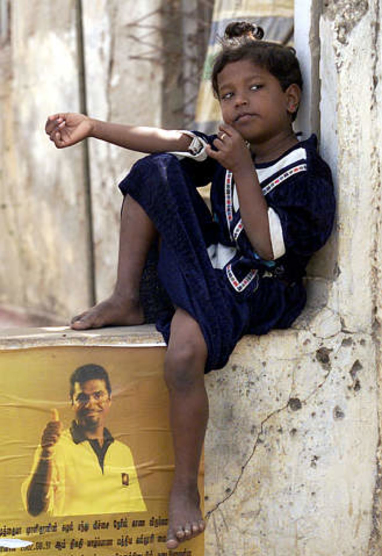 A Tamil girl sits and waits on a wall with bulletholes, for the start of a cricket match in the northern city of Jaffna, Sri Lanka on September 1, 2002. The poster on the wall anounces the arrival of Sri Lankan spin bowler Muttiah Muralitharan for a friendly cricket match at Jaffna Central College ground. Muralitharan and some other national Sri Lankan cricketers arrives in Jaffna after crossing Tamil Tiger rebel held Vanni area on Saturday to play a friendly cricket match against a Jaffna district side.