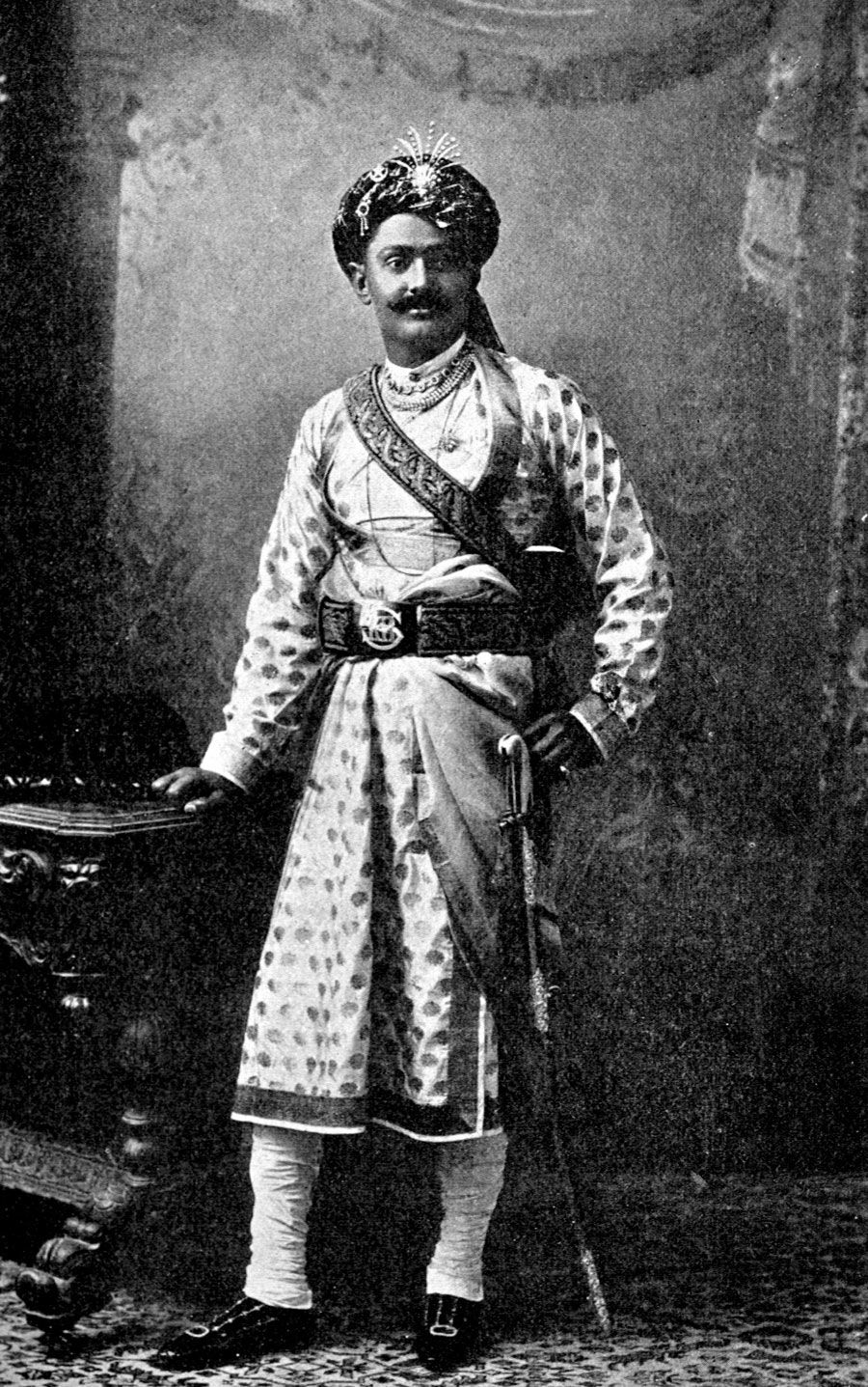Bonhams : BIKANER H.H. Ganga Singh, The Maharaja of Bikaner, standing full  length in ceremonial military costume with hand on sword, in a studio  setting with western-style carpet, red curtains and a